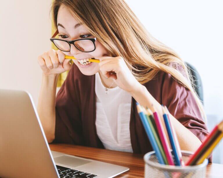 alt='Attractive Asian girl wearing glasses sits in front of a laptop and bites a yellow pencil.'