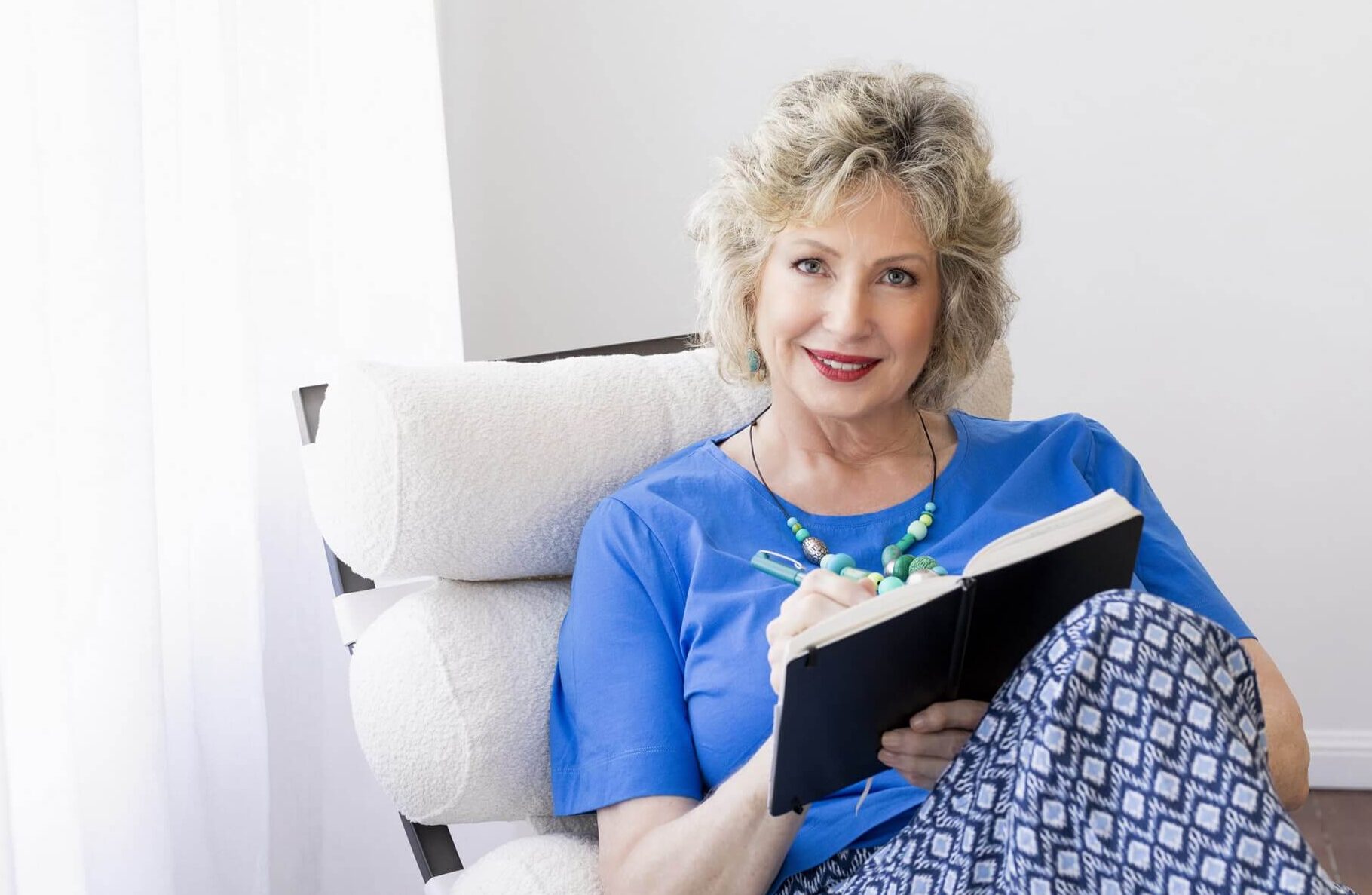 Blonde female wears light blue T-Shirt and blue patterned trousers. She sits on a modern white leather chair, writing in a book.