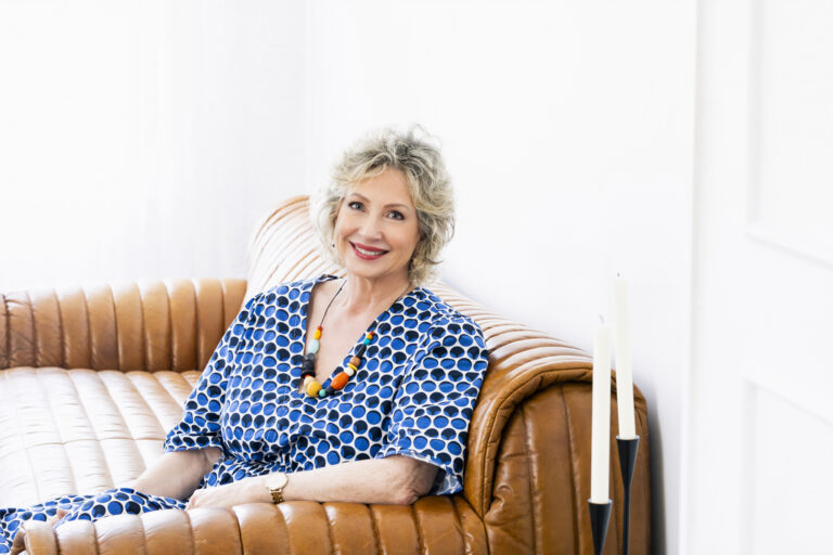 Photo of a blonde woman sitting on a tan leather couch. She wears a colourful necklace and a blue and white large spot dress. Background has white walls and white candles in the foreground.